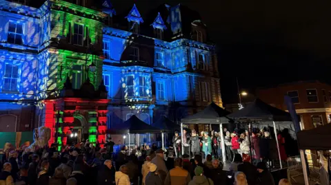 BBC A group of people stood around a stage which is positioned in front of an illuminated building.