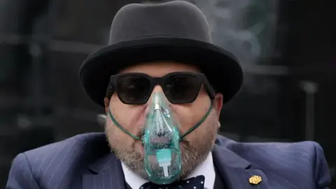 PA Media Nicholas Rossi, weaing a blue pin-stripe suit, bow tie and a black bowler hat. He is unshaven and is also wearing sunglasses and an oxygen mask.