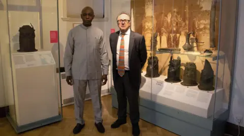 University of Cambridge Prince Aghatise Erediauwa (left) and Prof Nicholas Thomas (right) are photographed looking at the camera in a museum. Behind them are display cases with various sculptures.