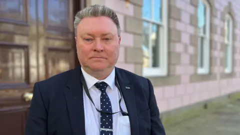 BBC A man wearing a suit stands on a pavement in front of a pink brick building. He is wearing a navy blazer, a white shirt and a blue and white spotty tie. 