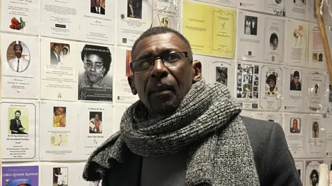 Jon Wright/BBC Max Thomas from Ipswich Windrush Society looks at the camera. He has a thick grey scarf around his neck and wears a dark suit jacket.