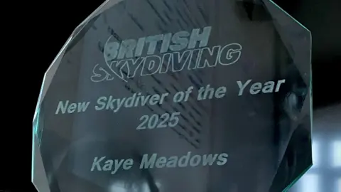 Stuart Hall A round transparent silver disc shaped trophy with jagged edges which says British Skydiving New Skydiver of the Year 2025 Kaye Meadows on it. 