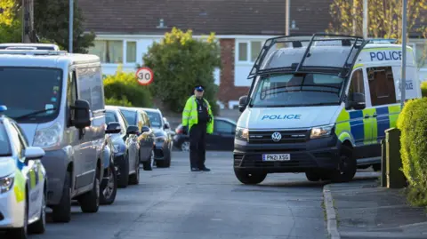 A police officer in a luminous reflective jacket stands in the middle of a residential street next to a large liveried police van with a riot shield above its windscreen. 