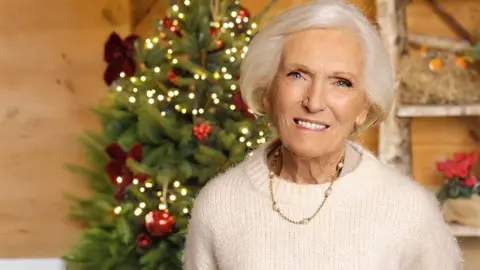 Mary Berry in front of a Christmas tree. She has white hair and wears a white jumper with a gold necklace