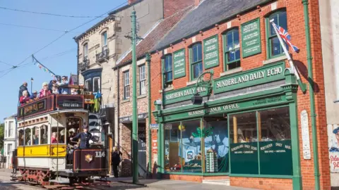 Getty Images View of a tram dating from Edwardian times with a number of people on board and a conductor in front of the spiral steps leading to the top deck. It is in front of a shop from the same era, Beamish Branch Office, with double fronted plate glass windows. Boards above the windows advertise the Northern Daily Mail and Sunderland Daily Echo