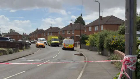 A residential street with police tape across the front. In the distance you can see several vehicles including an ambulance and a police van.