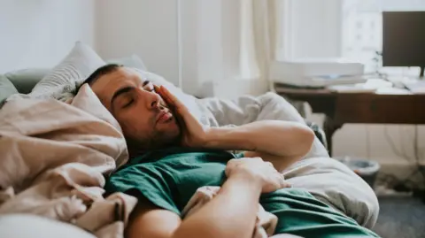 Getty Images A man wakes up on a sofa, surrounded by sheets and duvets and pillows. He rubs his eyes and face as he tries to rouse himself.