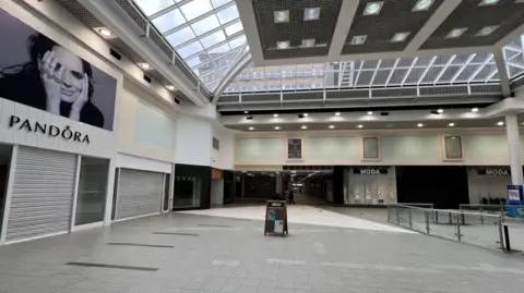 The picture shows the empty Nicholsons shopping centre with the shutters for businesses pulled down. A lone 'A' board stands in the middle of the shot with the centre's grey stone paved walkway devoid of shoppers. 