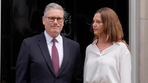 PA Media Sir Keir Starmer stood with his wife Victoria Starmer outside the door of No.10 Downing Street. The door is dark black. Starmer is wearing a suit, a white shirt, and a red tie. His wife is wearing a white shirt.