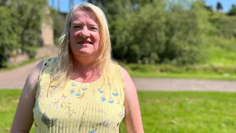 Catherine Ritchie looking straight at the camera. She has blonde hair past her shoulders. She is wearing a pale yellow, sleevless top with colourful birds on it. She is standing in front of a blurred background of greenery.