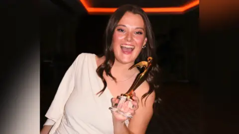 WIC/Louisa Rechenback A girl with dark brown hair and a cream dress holding a gold award.