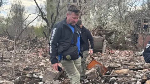 Nowzad Pen Farthing is wearing grey trousers, a blue T-shirt and black jumper. He is walking over a large pile of rubble.