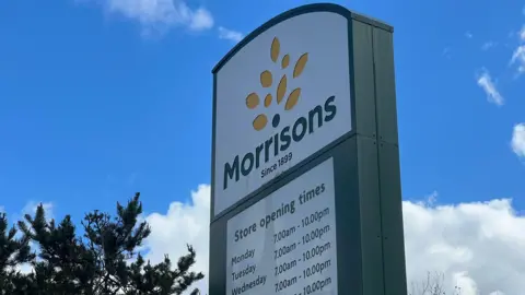 A tall Morrisons sign displaying the company logo and store opening times. The sign reads 'Morrisons - Since 1899' in dark green lettering. It stands against a bright blue sky with scattered clouds.