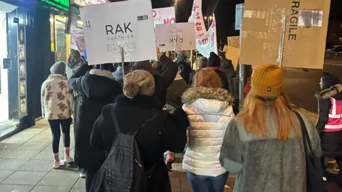 James Hill A back view of mostly women walking up a street in Northampton in 2024. They are holding up placards and are dressed for winter conditions with hats and coats 