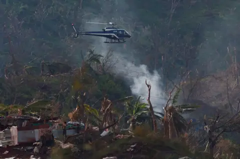 Gonzalo Fuentes/Reuters Sebuah helikopter dari Gendarmerie Prancis terbang di atas desa Longoni setelah Topan Chido di Mayotte. Pepohonan dan bangunan rata atau rusak parah dan asap mengepul dari tanah, lalu dua orang memeriksa kerusakan tersebut.