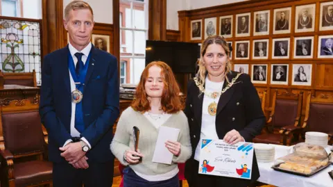 DOUGLAS CITY COUNCIL Evie, a teenager with ginger hair, with her eyes closed holding the award. She is standing between Douglas Mayor Natalie Byron-Teare, who is wearing her official chain, and consort Andy Teare in the council chamber.