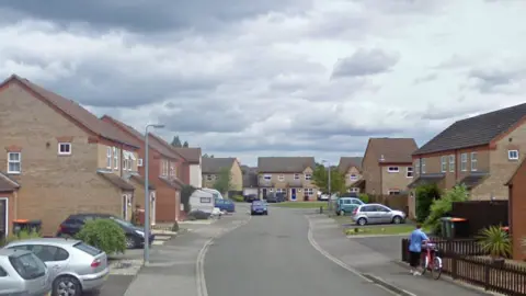 A general view of Fisher Close in Barton le Clay. Houses line the street and cars can be seen parked on driveways. A woman is pictured pushing a bike on the pavement.