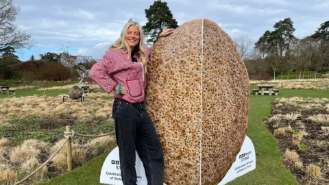 A woman wearing a red patterned jacket and black jeans is stood in an ornamental garden next to a card which is the same height as her. The card resembles a traditional Staffordshire oatcake.