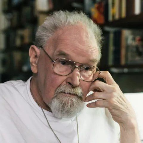 PA Media Alasdair Gray, a grey-haired man with a grey beard and moustache, stares solemnly to the side, his left hand at his cheek, wearing a plain white t-shirt and thin-rimmed glasses.