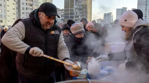 A man pours soup on a pastic cup as a crowd of people wearing winter clothes wait in line