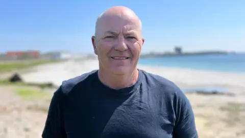 Donald Gillies stands with his back to a sandy coast. He is bald and wearing a navy blue t-shirt