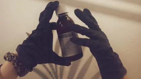 Royal Photographic Society/Ruby Hall A person holding a bottle of medicine with black gloves on