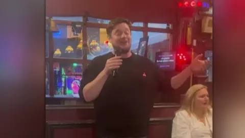 Man in black sweater closes his eyes as he sings into microphone - a woman in a white shirt with blonde hair sits behind him. You can see the words on karaoke machine.