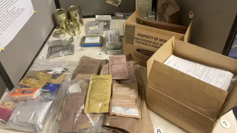 Packets of rations in a pile on a table, with gold tins of rations behind. There are cardboard boxes on the table that say property of MOD