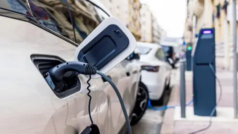 Getty Images A white electric vehicle parked on the side of the road as it charges. The dark grey charging station is on the footpath. There is another white car and a black vehicle parked in front.