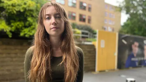 Julia Gregory/BBC A woman with long red hair, wearing a dark green top. She is standing outside a university campus.