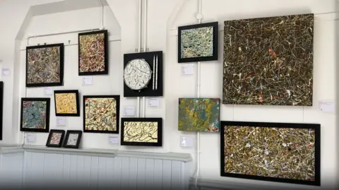 Pieces of art in all different shapes hang on a white wall. They are all different and have a black border around them. 