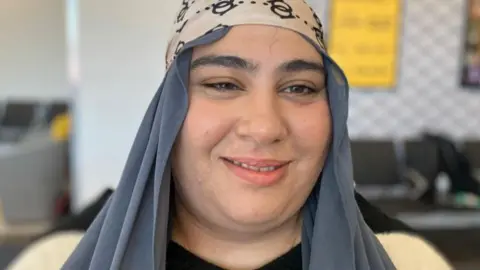 Andrew Sinclair/BBC Angelina Aziz is wearing a grey or navy head scarf and is standing inside.