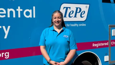 A woman smiling at the camera, slightly squinting at the sun. She's in a blue polo shirt. Behind her is the blue of the side of the van, with the TePe logo next to her head.