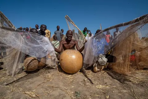 Sani Maikatanga A bare-chested fisherman sits with his gourd and nets on dusty ground. 