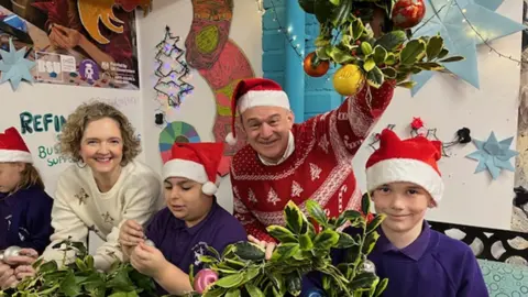 Ed Davey wearing a red and white Christmas jumper with trees on and a Santa hat, standing behind two boys in purple polo shirts, and also in Santa hats, with a young female teacher. Ed is holding up a Christmas decoration with holly and baubles and is smiling to camera