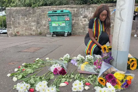 A woman, wearing a scarf featuring the colours of the Zimbabwe flag, crouches to lay flowers at the foot of a lampost on a pavement near the murder scene