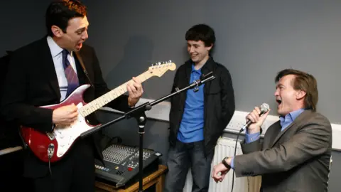 PA Media Andy Burnham on a red electric guitar to the left of the picture. He is wearing a suit and his mouth is open as though he is singing. Feargal Sharkey is to the right of the picture and is singing into a microphone. There is a young boy standing between them, smiling.