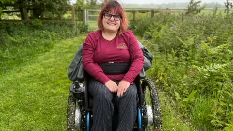 A young woman with red and brown hair smiles in to the camera. She is seated in a wheelchair is a wearing a dark red long-sleeved top and dark trousers with white trainers.