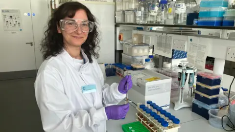 Amy Holmes/BBC A picture of a dark haired woman, wearing safety goggles and a lab coat, in a laboratory, testing samples 