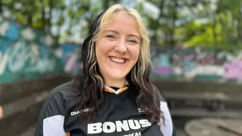 A woman with dark hair and blonde highlights is smiling as she looks at the camera. She is wearing a black Hull City shirt and is in an outside setting with wooden tables and trees in the background.