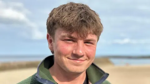 BBC Euan smiling at the camera, with a yellow beach and blue sea behind them. He has brown hair and is wearing a green fleece.