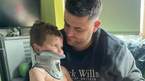 A boy wearing a neck brace is sitting on his father's knee in a room in a hospital.
