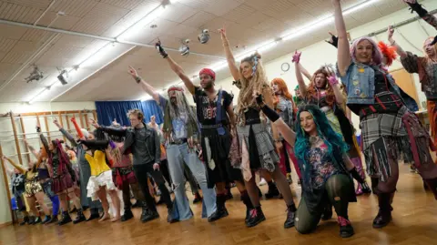 Hannah Ogden A group of people in a variety of costumes rehearse in a room with a wooden floor. 