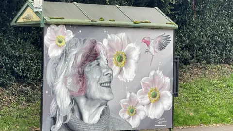 Nue/Sam Art A bus shelter with a painting of a woman with grey hair with pink at the front who is smiling and surrounded by four white and pink flowers with yellow and green centres and a bird. 