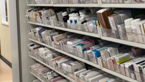 Rows of prescription drugs within a pharmacy. Five rows of the prescription draws can be seen within the picture.