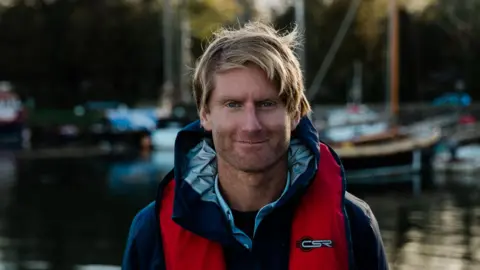 A photo of Chris Ranford, his head and shoulders. He has blond hair and blue eyes and is wearing a blue and red jacket. There is a boat on the water in the blurred background behind him.