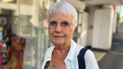 Joan Kelly is smiling at the camera as she stands in a street. She has short white hair and is wearing a pair of glasses. She is wearing a white shirt over a flowery top and a bag is slung over her shoulder. She is also wearing make-up and a silver necklace with a green stone pendant. The background is blurred behind her.