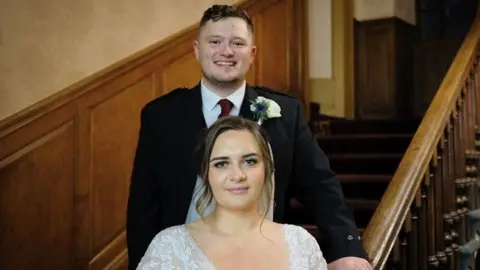 Weronika Somerville pose couples on a ladder in the wedding site, which has wood-paneal walls and banists. Cameron stands behind her bride - she is wearing a kilt, jacket, burgundy tie and a flower in her buttonhole, while Veronica is in a lace gown and veil.