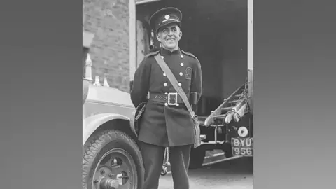 A man posing for the camera in a button up jacket and hat of the firefighter's uniform. He is smiling at the camera and behind him there is an old vehicle with a ladder on the back of it.