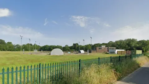 Google A green field behind a dark green iron fence. A brown brick building and a old cricket pitch and floodlights are in the middle of the picture. 